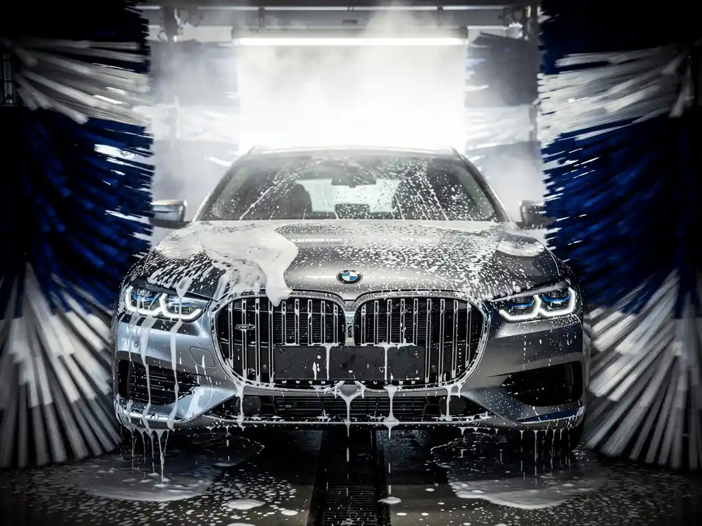 Glanzende auto in automatische carwash tunnel, omgeven door blauwe en witte textielborstelgordijnen met schuim en waterdruppels.